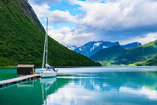 A white sailboat is docked at a wooden pier, surrounded by the stunning beauty of the Norwegian fjords. Lush green hills rise on the side of the calm, clear waters Norangsfjorden Norangdal Urke Norway