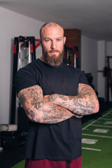 strong sportsman with beard and tattoos standing with crossed arms in gym and looking at camera,...