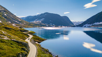 A single car winds its way through the majestic Norwegian landscape, a stunning lake reflecting the sky and snow-capped peaks. Langvatnet, Geiranger, Norway © Fokke Baarssen