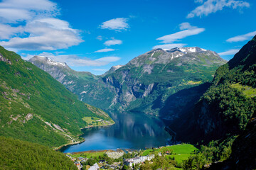 A scenic view of a Norwegian fjord, with lush green mountains surrounding a calm blue body of water. The sky is clear and blue, with fluffy white clouds. Geiranger Fjord Norway in summer