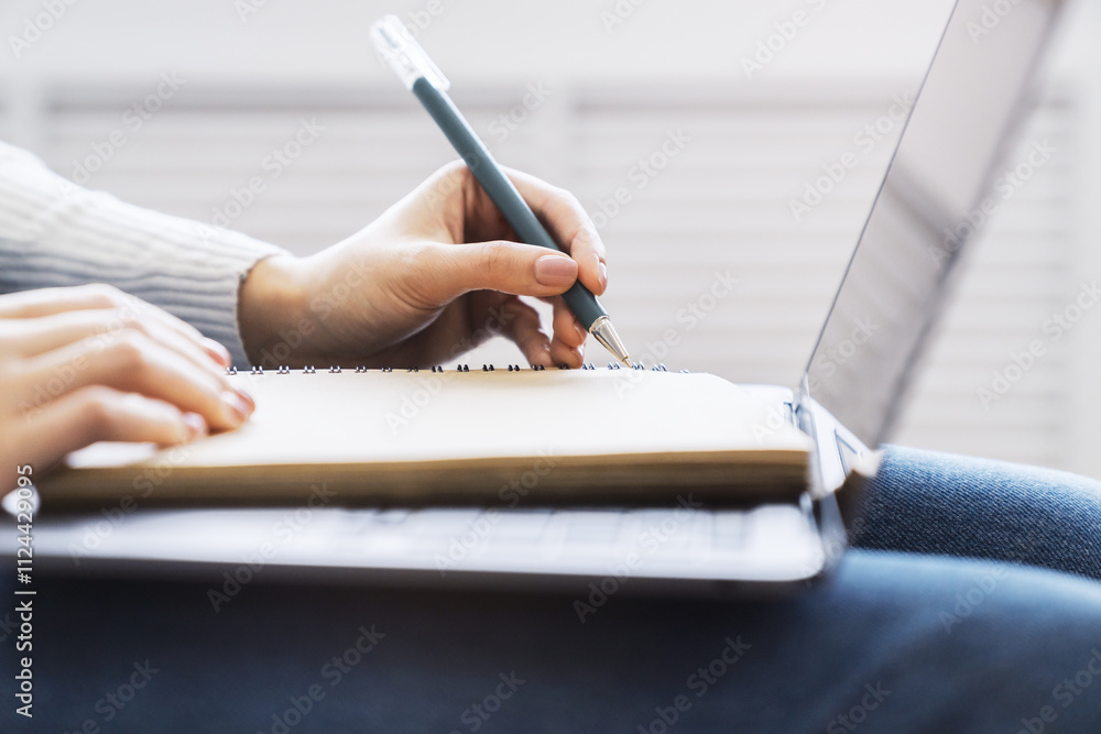 Poster close-up depiction of a woman's hand taking notes in a notepad on a sleek laptop, with a blurred bac