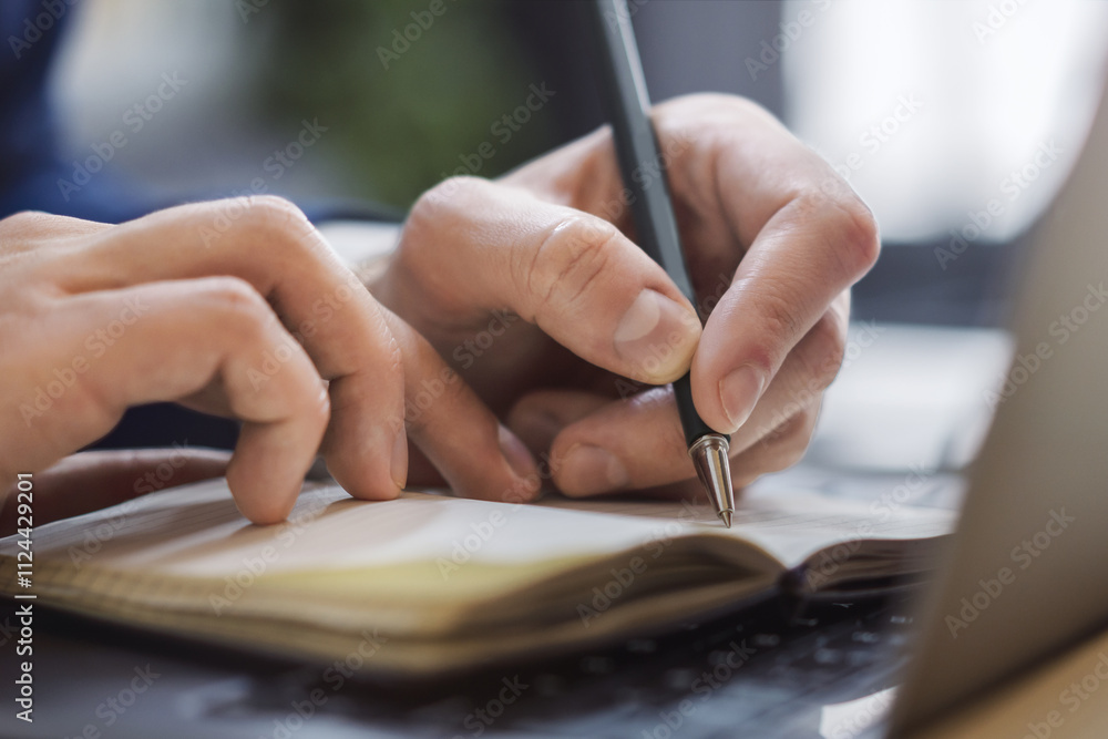 Poster Detailed view of a man's hand jotting down notes in a notepad on a modern laptop, with a blurred background