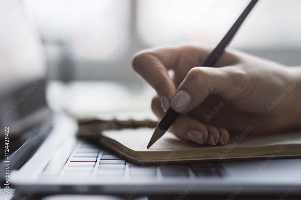 Poster The image captures a woman's hand in close view as it writes in a notepad on a chic laptop, background out of focus