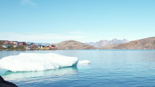 An iceberg (ice floe) floating on calm Arctic waters near a picturesque, traditional, and remote small village in Greenland.
