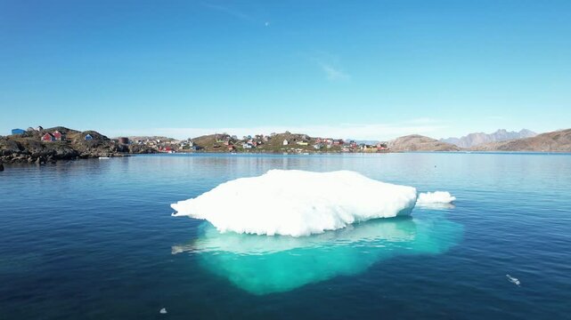 Aerial footage of an iceberg (ice floe) floating on calm Arctic waters near a picturesque, traditional, and remote small village in Greenland. Filmed with a Drone.