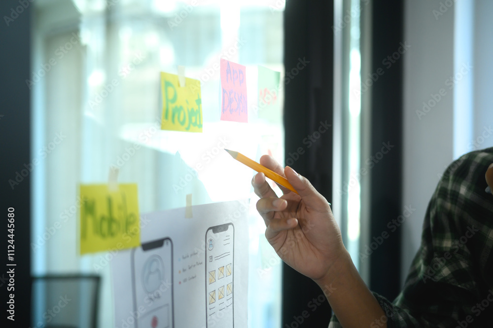 Poster close up of team member pointing at colorful sticky notes and app wireframe sketches on a glass wall