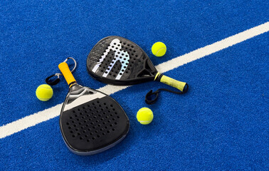 A padel or paddle tennis racket and a bright yellow tennis ball lie on a blue court. The net and a blurred figure of a player are visible in the background. © Angelov