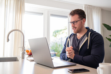 working at home in the kitchen. caucasian male sitting at the kitchen counter behind his laptop working and communicating. Dressed in a blue hoodie, wearing glasses.