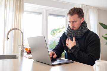 working at home in the kitchen. caucasian male sitting at the kitchen counter behind his laptop working and communicating. Making a difficult looking face, sick nog well, wearing a scarf.