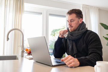 working at home in the kitchen. caucasian male sitting at the kitchen counter behind his laptop working and communicating. Wearing a scarf and not feeling well, sick holding tissue in the hand