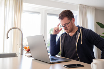 working at home in the kitchen. caucasian male sitting at the kitchen counter behind his laptop working and communicating. Dressed in a hoodie, wearing glasses.