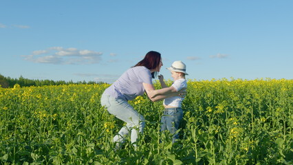 Fototapeta premium Woman And Their Son Are Playing In A Field Of Rapeseed. Happy Child Playing Joyfully In Fields Of Blooming Yellow Rapeseed.