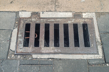 A close up of a metal storm drain cover on a sidewalk, featuring rectangular slots and surrounding...