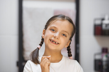 Portrait of cheerful Caucasian girl showing freshly fallen tooth. She smiles while looking in bathroom mirror. Youthful pride and childhood milestone captured.