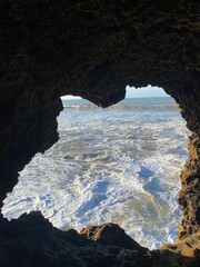 A stunning heart-shaped cave opening framing a view of the ocean waves crashing below.