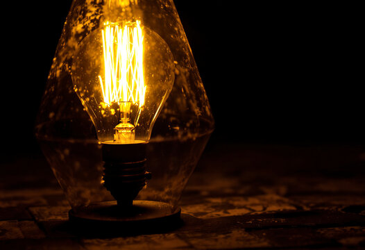 A vintage glass lamp with a glowing incandescent light bulb inside, casting a warm, amber-colored glow in a dark room