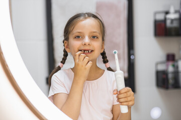 Little Caucasian girl with missing baby teeth using electric toothbrush and smiling in bathroom mirror, showing confidence and dental care.