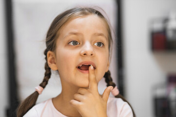 Little Caucasian girl with fallen milk teeth touches loose tooth while looking in mirror in bathroom. Moment captures curiosity and childhood growth.