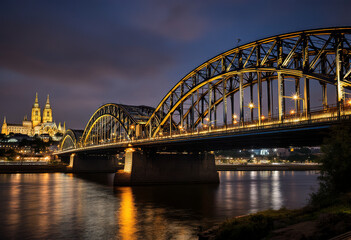 Fototapeta premium A large illuminated bridge with arched steel structures spanning a river at night, with a cathedral-like building visible in the background