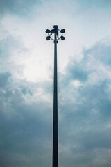 A tall street light pole with multiple floodlights against a cloudy sky, creating a dramatic and moody atmosphere