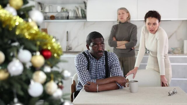 Upset middle-aged man sitting at table while two women quarreling to him standing behind in the kitchen decorated with Xmas-tree. High quality 4k footage