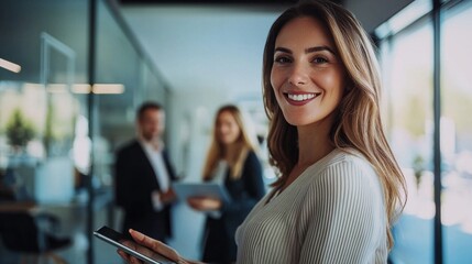 Fototapeta premium In a bright, modern office, a smiling business leader holds a tablet, radiating confidence and approachability as she interacts with her colleagues in the background