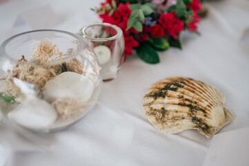 Eine maritime Tischdekoration mit einer Muschelschale, einem Glasgefäß mit Dekoelementen wie Steinen und Stroh sowie einer kleinen Kerze. Im Hintergrund ist ein Blumenarrangement zu sehen. 