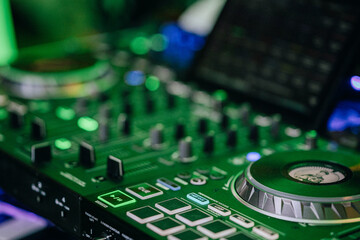 Ein professionelles DJ-Mischpult mit verschiedenen Reglern, Tasten und Jogwheels, beleuchtet in grünen Tönen. Im Hintergrund sind verschwommene Details eines Laptops und weitere DJ-Equipment sichtbar.