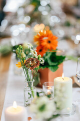 Eine stilvolle Tischdekoration mit Kerzen, kleinen Blumenvasen und orangefarbenen sowie weißen Blüten. 