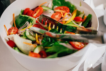 Ein frischer Gemüsesalat in einer weißen Schüssel, dekoriert mit bunten Zutaten wie Tomaten, Gurken, Paprika, roten Zwiebeln und Salatblättern. Eine Salatzange liegt bereit, um den Salat zu servieren.