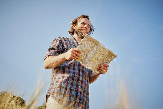 Smiling young man holding map in field under blue sky