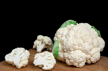 Fresh cauliflower arranged on a wooden cutting board with separate florets on black background