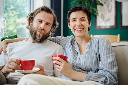 Young smiling couple sitting with cup of tea on sofa at home - Powered by Adobe