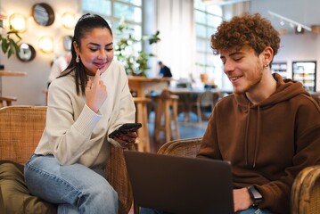 A woman holding a smartphone and a man in a brown hoodie smiling and interacting in a comfortable indoor area with vibrant decor and large windows in the background