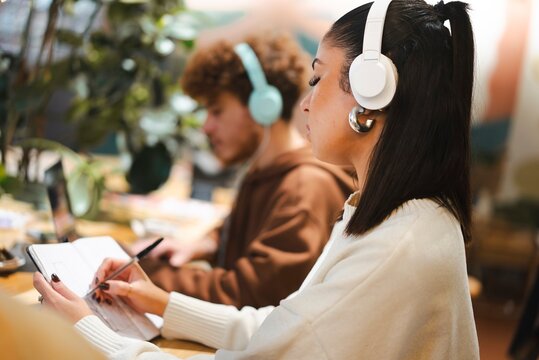A young woman in white headphones taking notes in a notebook while a man in teal headphones works on a laptop in the background, set in a cozy and bright cafe environment