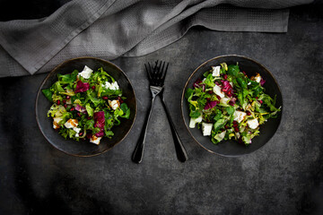 Mixed green salad with feta and pomegranate seeds, balsamico, and olive oil in black bowls on a dark table.