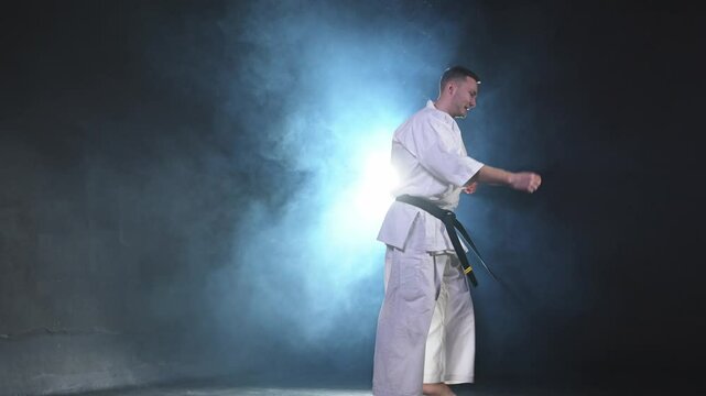 Strong man fighter in white kimono for karate martial arts with black belt jump kicking in black background. Man training moves practicing shadow fight preparing for fight training ready for sparring