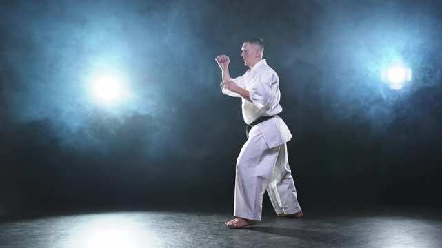 Strong man fighter in white kimono for karate martial arts with black belt preparing for fight training in black background. Man training moves kicks practicing shadow fight ready for sparring
