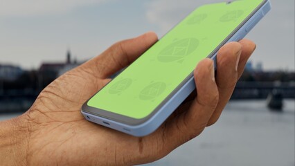 GREEN SCREEN CHROMA KEY Hand holding a mobile phone by a scenic river in an urban area, perfect for app mockup with cityscape background