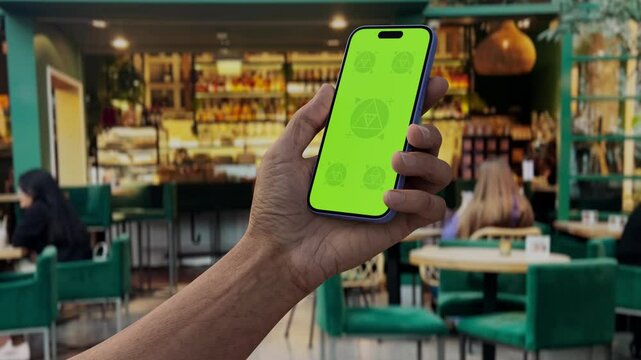 GREEN SCREEN CHROMA KEY Hand holding smartphone with green screen in a cozy cafe, ideal for app mockups and branding in a lively restaurant environment