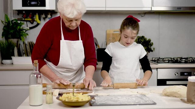 Beautiful little girl does good job with rolling pin rolling out piece of dough. Grannys cooking lessons are going positively. Grandmother and granddaughter preparing dough in cozy kitchen. Family