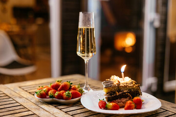 gourmet cake adorned with a candle rests on a plate beside a bowl of fresh strawberries, paired with a sparkling drink, all set on a rustic wooden table outdoors in the evening