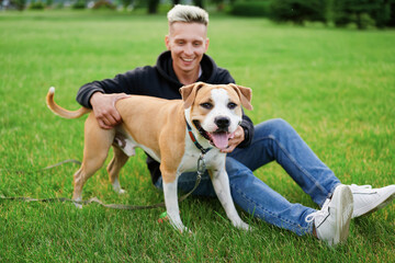 The dog is standing on the grass and looking into the frame with a smile and tongue out, next to the owner who is hugging it. American bulldog. Dog wearing a collar and leash.