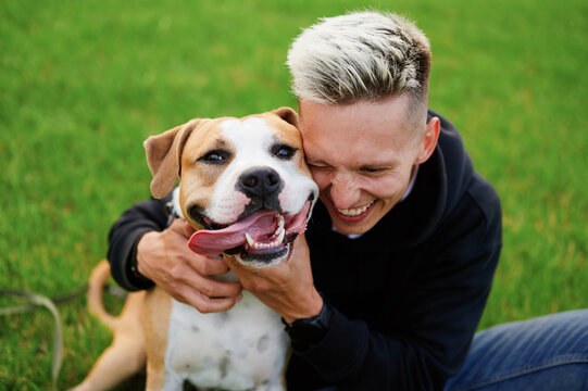 The dog smiles with his mouth open and tongue out in the arms of his breeder. A young caucasian man hugs his dog sitting on the grass in the park. American bulldog. Friendship of a dog and a man.