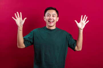 A cheerful young Asian man in a green T-shirt smiles brightly while raising one hand in a friendly wave, with the other hand on his hip, standing confidently against a bold red background