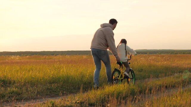 Parent teaches child to ride bicycle. Father and daughter in field at epic sunset. Child safety, dad regulates and controls beloved girl kid on bike, warning against injuries and bruises. Slow motion
