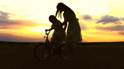 Fototapeta premium happy family. mother teaches little girl daughter ride bike sunset. concept happy family park. mom with child walks with bike park sunset sky. parent walks with kid together. happy childhood. girl