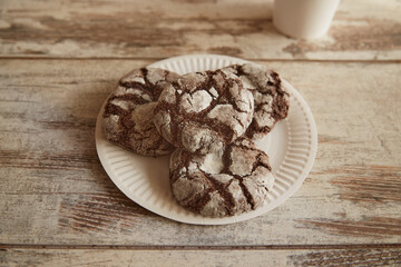 A full paper plate with chocolate cookies with cracks on an old rustic wooden table and a cup of coffee nearby