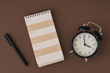 Planner pad, alarm clock and pen on brown background. Planner for organization and goal priorities. Writing down tasks and schedule reminders on office desk. Trendy brown color flat lay.