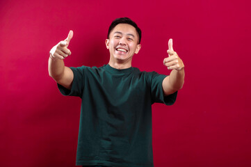 An Asian man in a green t-shirt smiles brightly, pointing directly at the camera with both hands,...